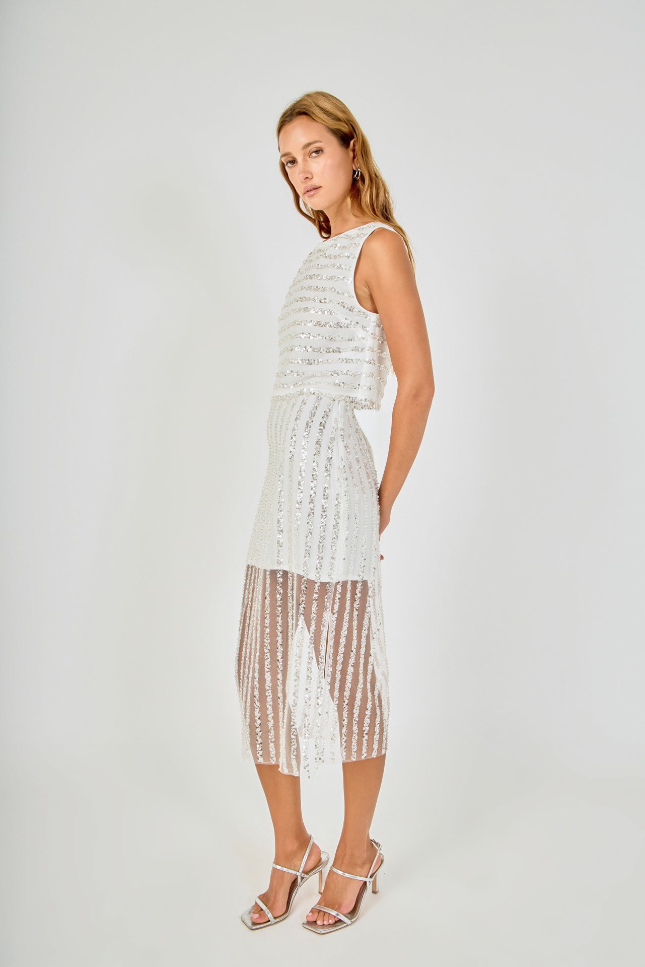 Side posed view of an off-white sequin stripe mesh midi skirt featuring an opaque mini skirt lining and sheer hem, styled with a matching sequin crop top.