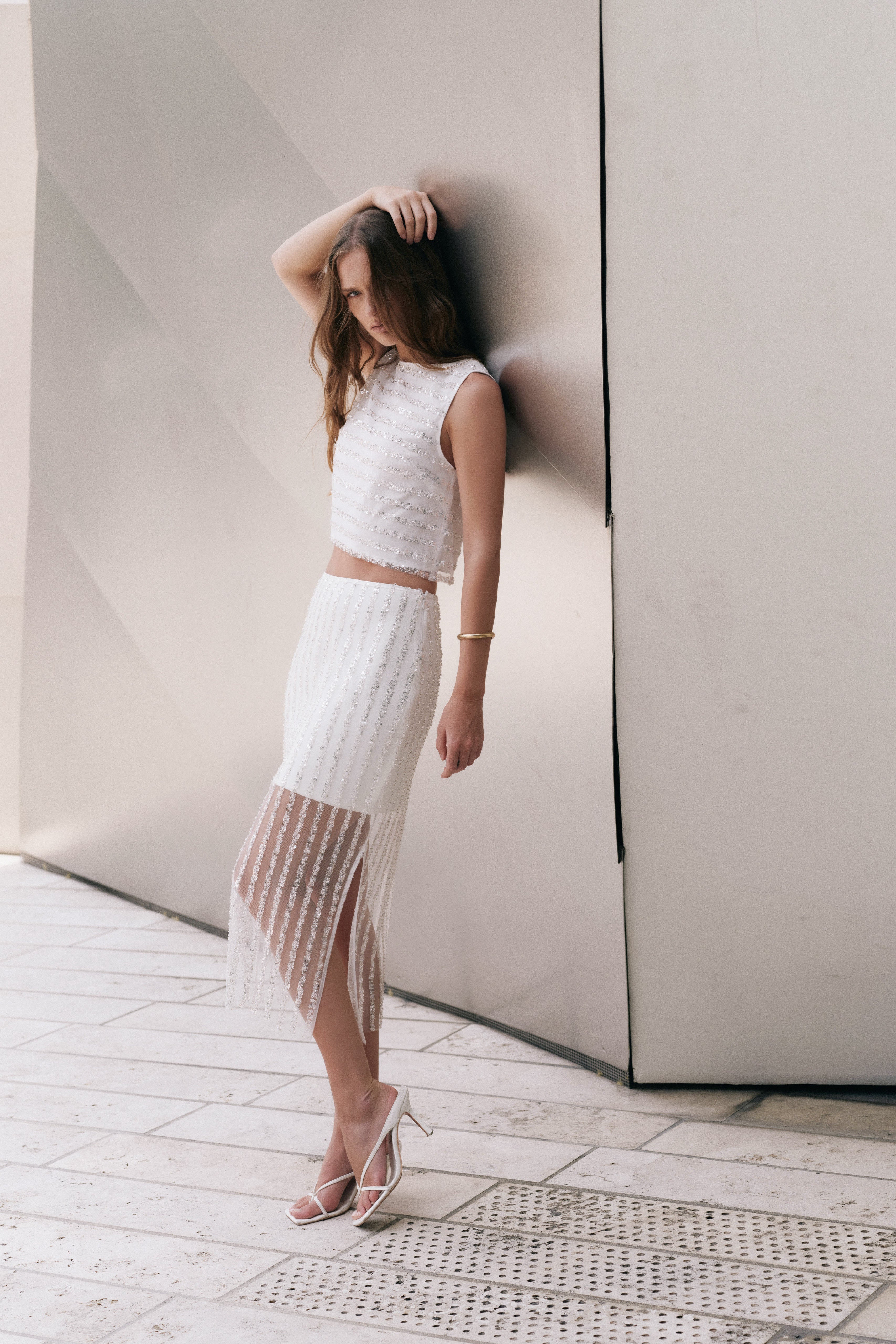 Full-length side profile view of an off-white high-waisted midi skirt featuring vertical sequin stripes, an opaque mini lining, a sheer lower hem, and a side slit.