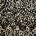 Detail view of black lace fabric featuring intricate scalloped embroidery, black bead embellishments, and a contrast nude lining.