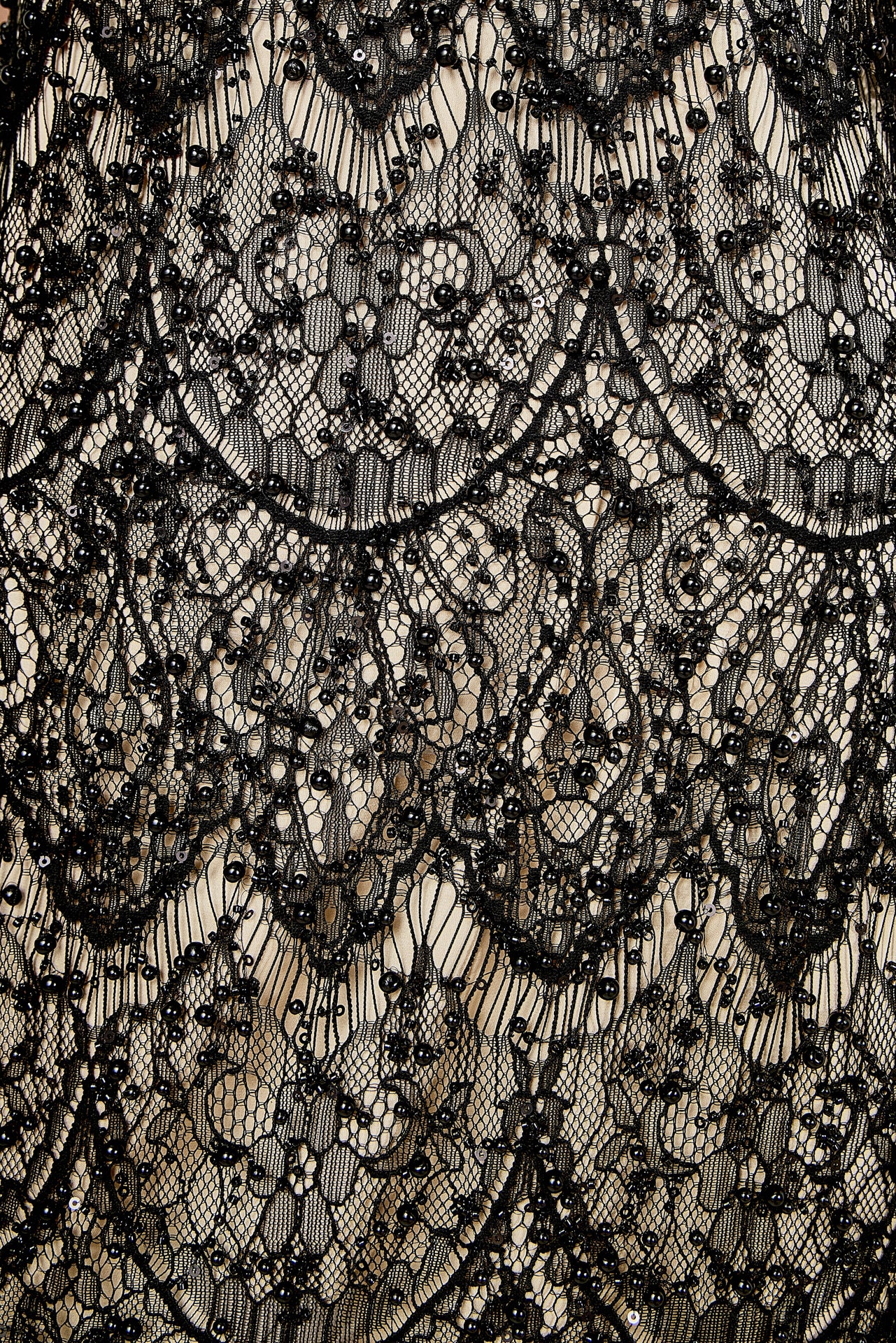 Detail view of black lace fabric featuring intricate scalloped embroidery, black bead embellishments, and a contrast nude lining.