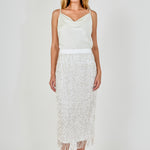 Full-length front view of a white high-waisted midi skirt with tiered sequin fringe and a fringed hemline, styled with a satin cowl-neck top.