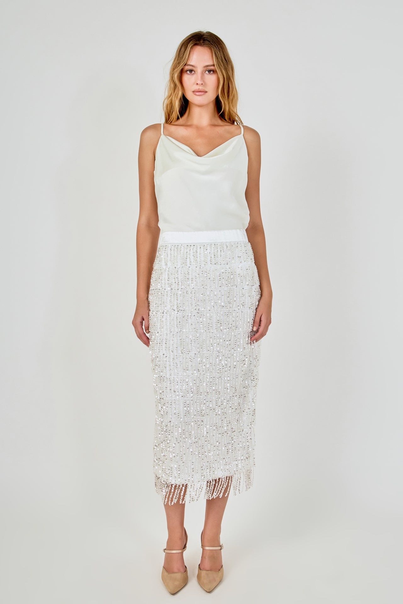 Full-length front view of a white high-waisted midi skirt with tiered sequin fringe and a fringed hemline, styled with a satin cowl-neck top.