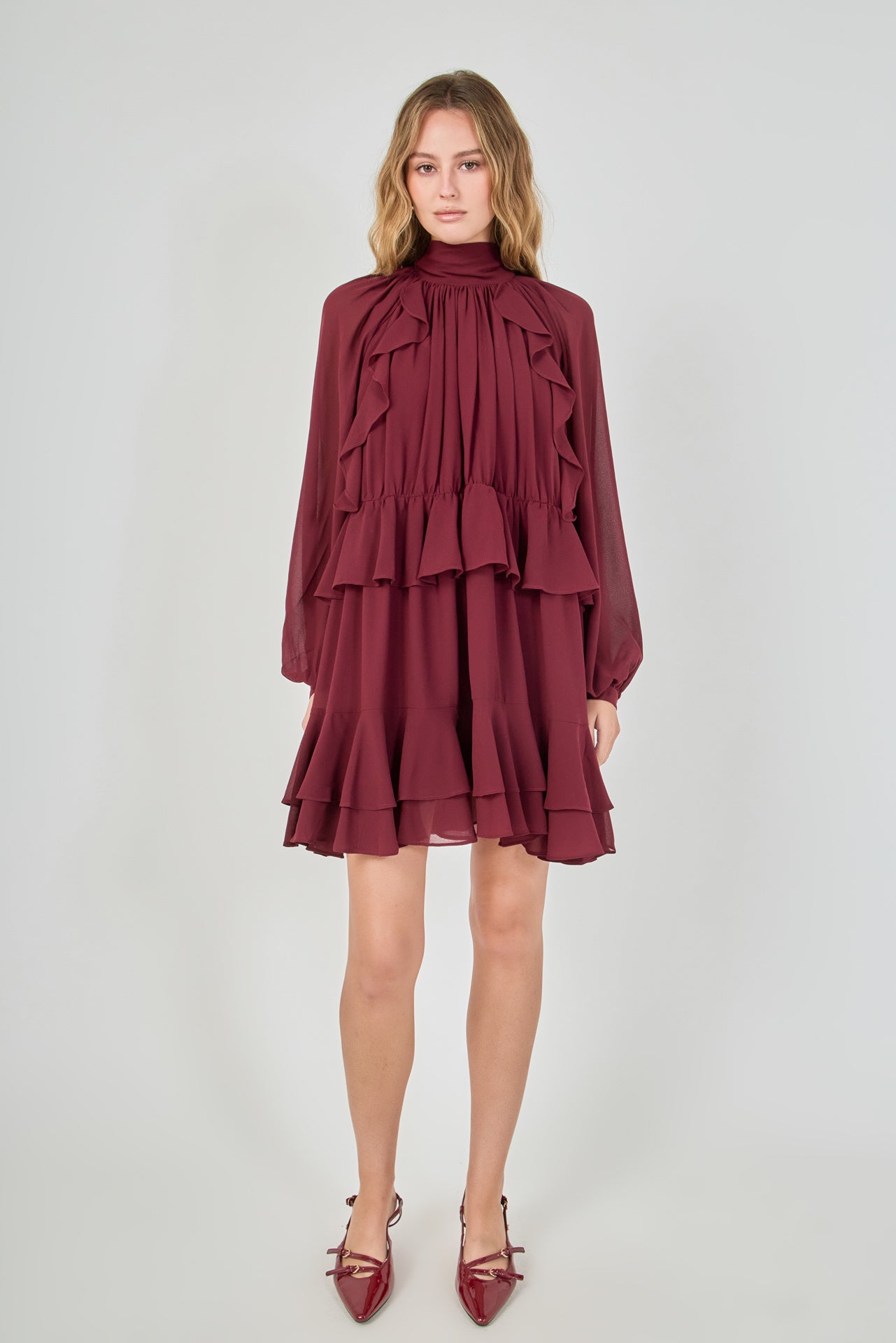 Full-length front view of a deep burgundy chiffon mini dress, featuring a smocked high neck, voluminous bishop sleeves, and a cascading tiered ruffled skirt.