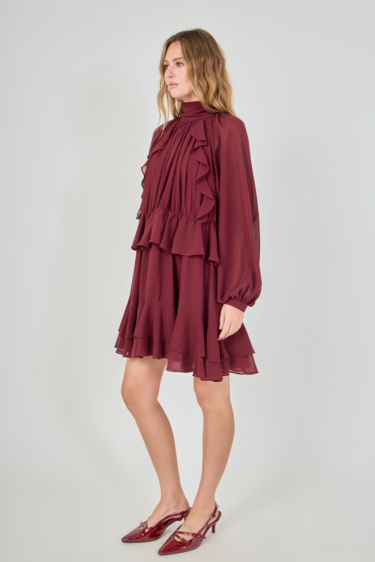 Profile view of the burgundy chiffon mini dress, showcasing the side silhouette, the fullness of the sheer balloon sleeves, and how the layers of the tiered ruffled skirt drape from the side.