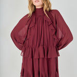 Mid-shot face-forward posed view of the deep burgundy chiffon mini dress. The model looks at the camera with hands placed on her hips, pulling the fabric slightly to define the waist and accentuate the ruffles