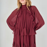 Mid-shot face-forward posed view of the deep burgundy chiffon mini dress. The model looks at the camera with hands placed on her hips, pulling the fabric slightly to define the waist and accentuate the ruffles