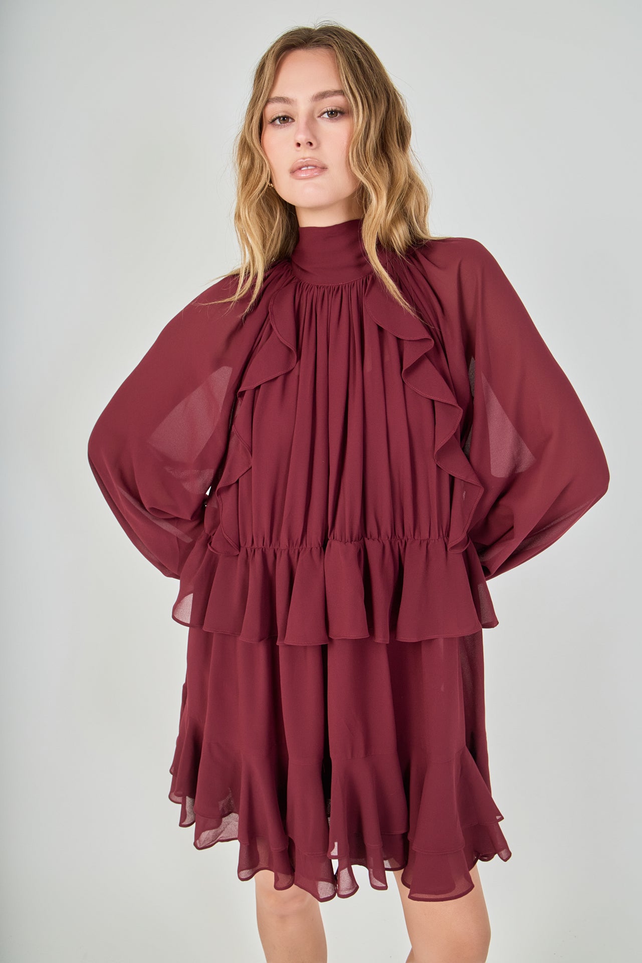 Mid-shot face-forward posed view of the deep burgundy chiffon mini dress. The model looks at the camera with hands placed on her hips, pulling the fabric slightly to define the waist and accentuate the ruffles
