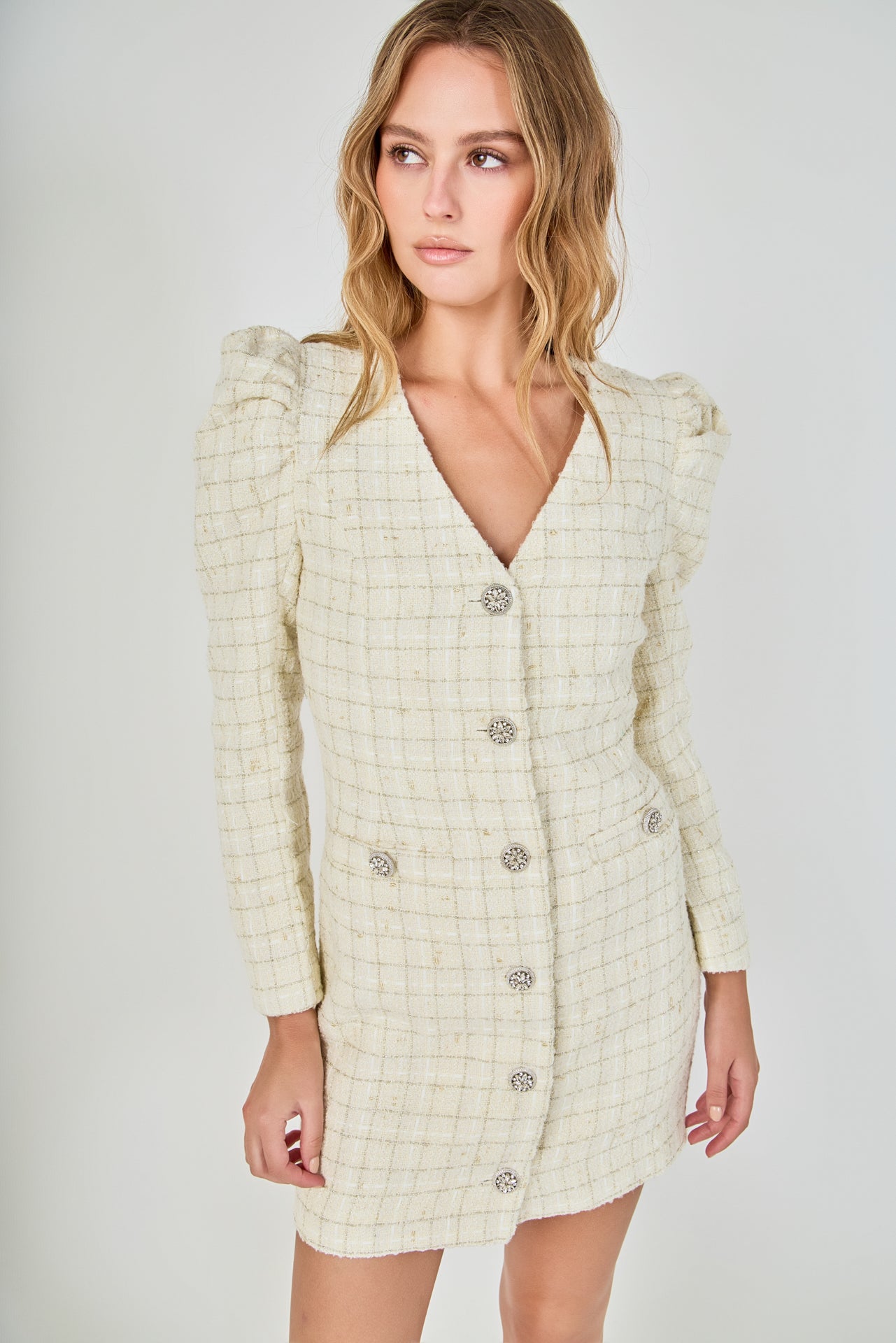 Face-forward posed view of an ivory long-sleeve tweed blazer mini dress featuring a deep V-neckline, structured puff shoulders, and crystal button closures.