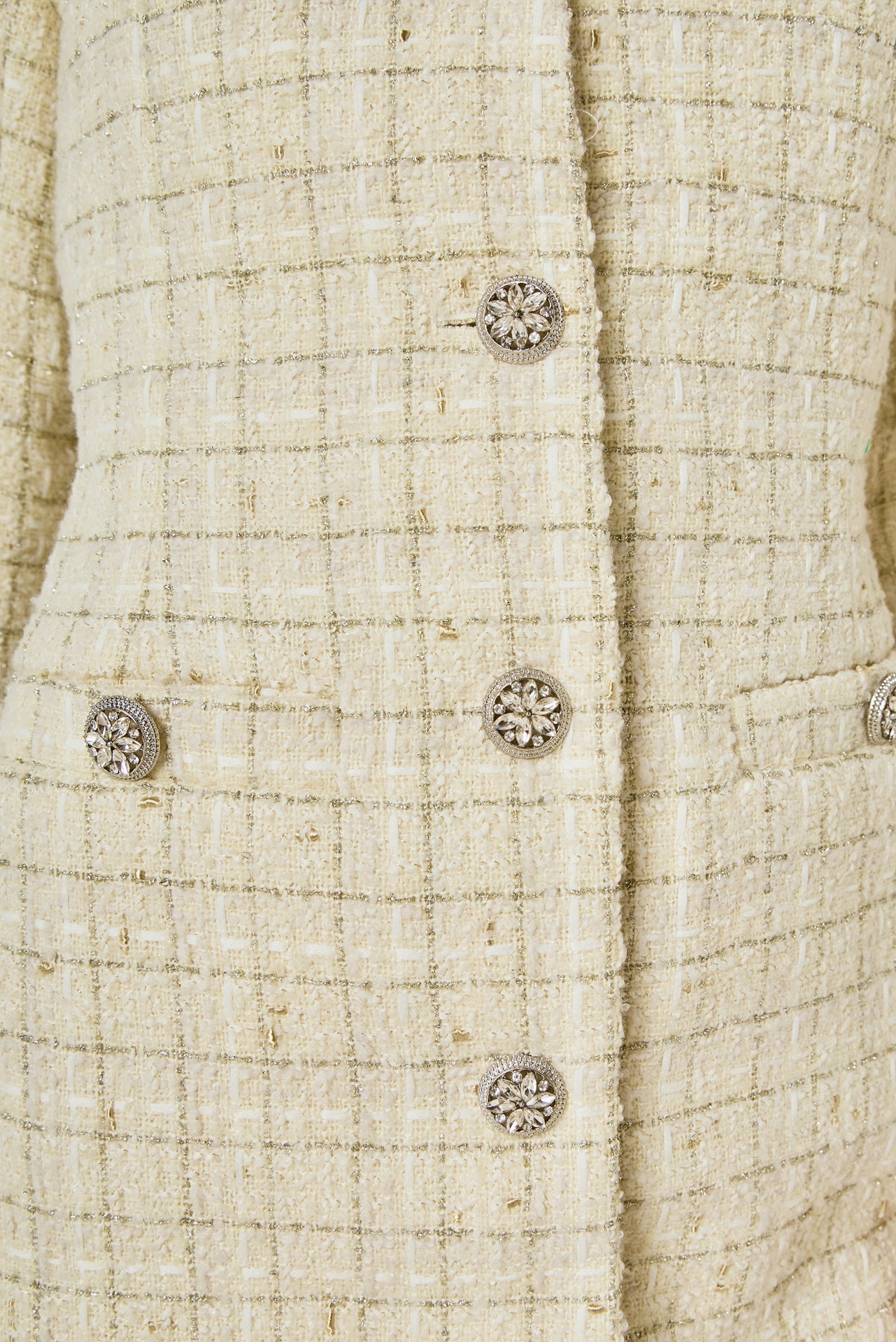 Ultra close-up detail view of ivory woven tweed fabric featuring a subtle gold metallic windowpane grid, ornate crystal floral buttons, and faux flap pockets.