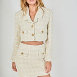 Face-forward posed view of an ivory cropped tweed jacket and matching high-waisted tweed mini skirt set featuring textured gold button accents.