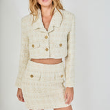 Face-forward posed view of an ivory cropped tweed jacket and matching high-waisted tweed mini skirt set featuring textured gold button accents.