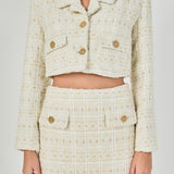Close-up view of an ivory cropped tweed jacket and matching mini skirt set highlighting the wide notch collar, front flap pockets, and textured gold buttons.