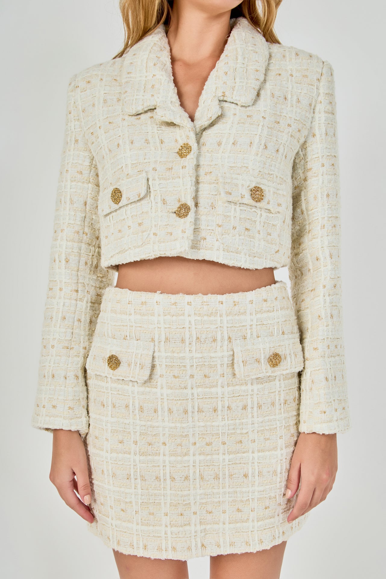 Close-up view of an ivory cropped tweed jacket and matching mini skirt set highlighting the wide notch collar, front flap pockets, and textured gold buttons.