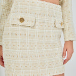 Close-up view of an ivory high-waisted tweed mini skirt highlighting the textured windowpane weave and front flap accents with decorative gold buttons.
