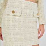 Close-up view of an ivory high-waisted tweed mini skirt highlighting the textured windowpane weave and front flap accents with decorative gold buttons.