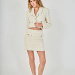 Face-forward posed view of an ivory high-waisted tweed mini skirt and matching cropped jacket set featuring decorative gold buttons.