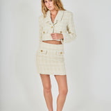Face-forward posed view of an ivory high-waisted tweed mini skirt and matching cropped jacket set featuring decorative gold buttons.