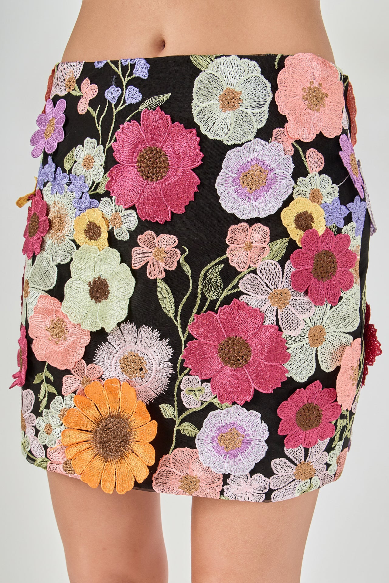 Detail view of black skirt fabric featuring dense multi-colored floral embroidery in vibrant shades of pink, orange, yellow, mint green, and lavender.