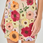 Detail view of beige skirt fabric featuring dense multi-colored floral embroidery in shades of red, orange, yellow, pink, and purple with green stems.
