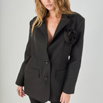 Dynamic, angled front view of a model looking forward, showcasing the black blazer with its 3D rosette detail, paired with black capri pants and black heels.