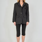 Full-length front view of a model wearing a black tailored blazer featuring a prominent 3D rose appliqué, two-button closure, and flap pockets, paired with fitted black capri pants with split hems.