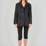 Full-length front view of a model wearing a black tailored blazer featuring a prominent 3D rose appliqué, two-button closure, and flap pockets, paired with fitted black capri pants with split hems.