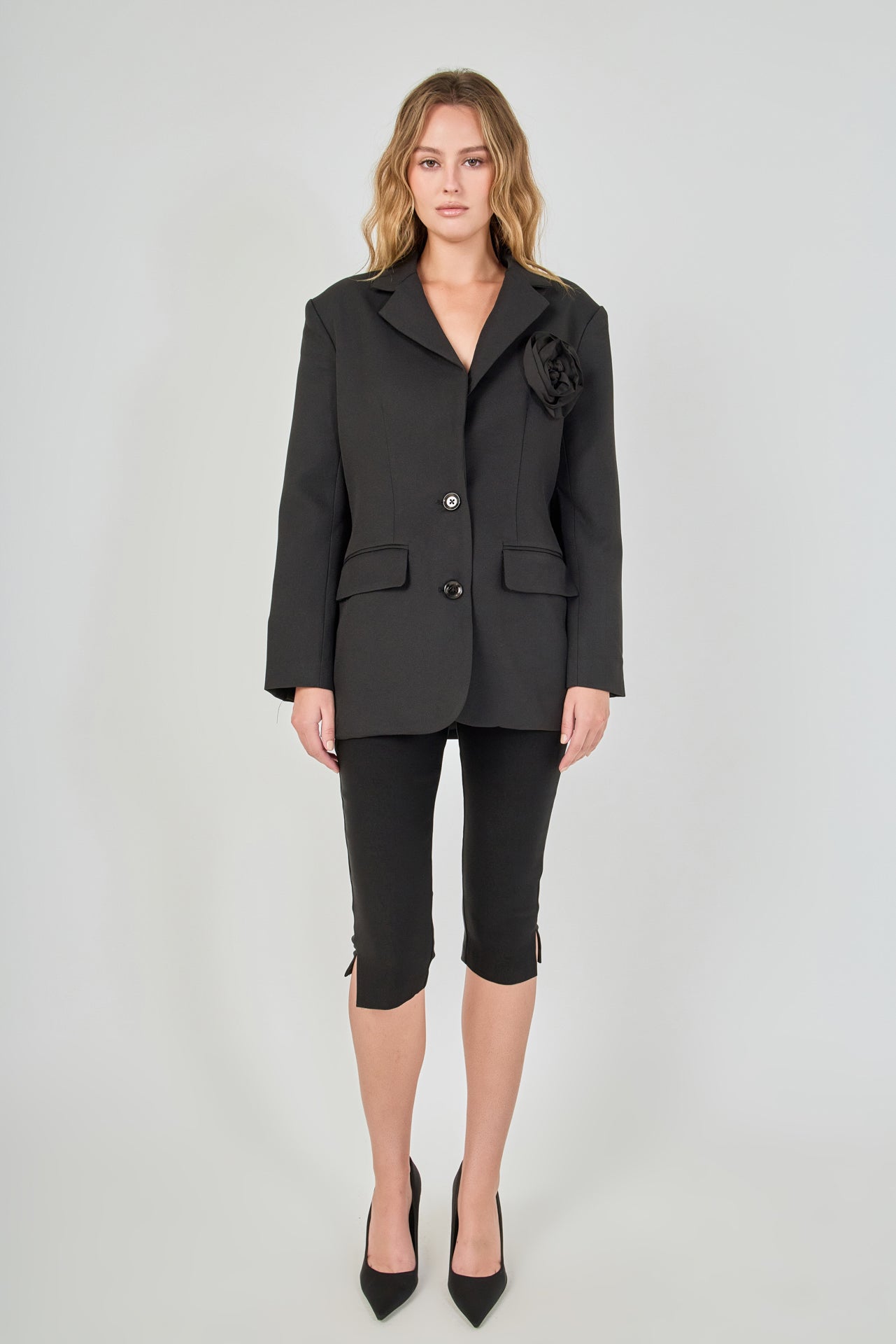 Full-length front view of a model wearing a black tailored blazer featuring a prominent 3D rose appliqué, two-button closure, and flap pockets, paired with fitted black capri pants with split hems.