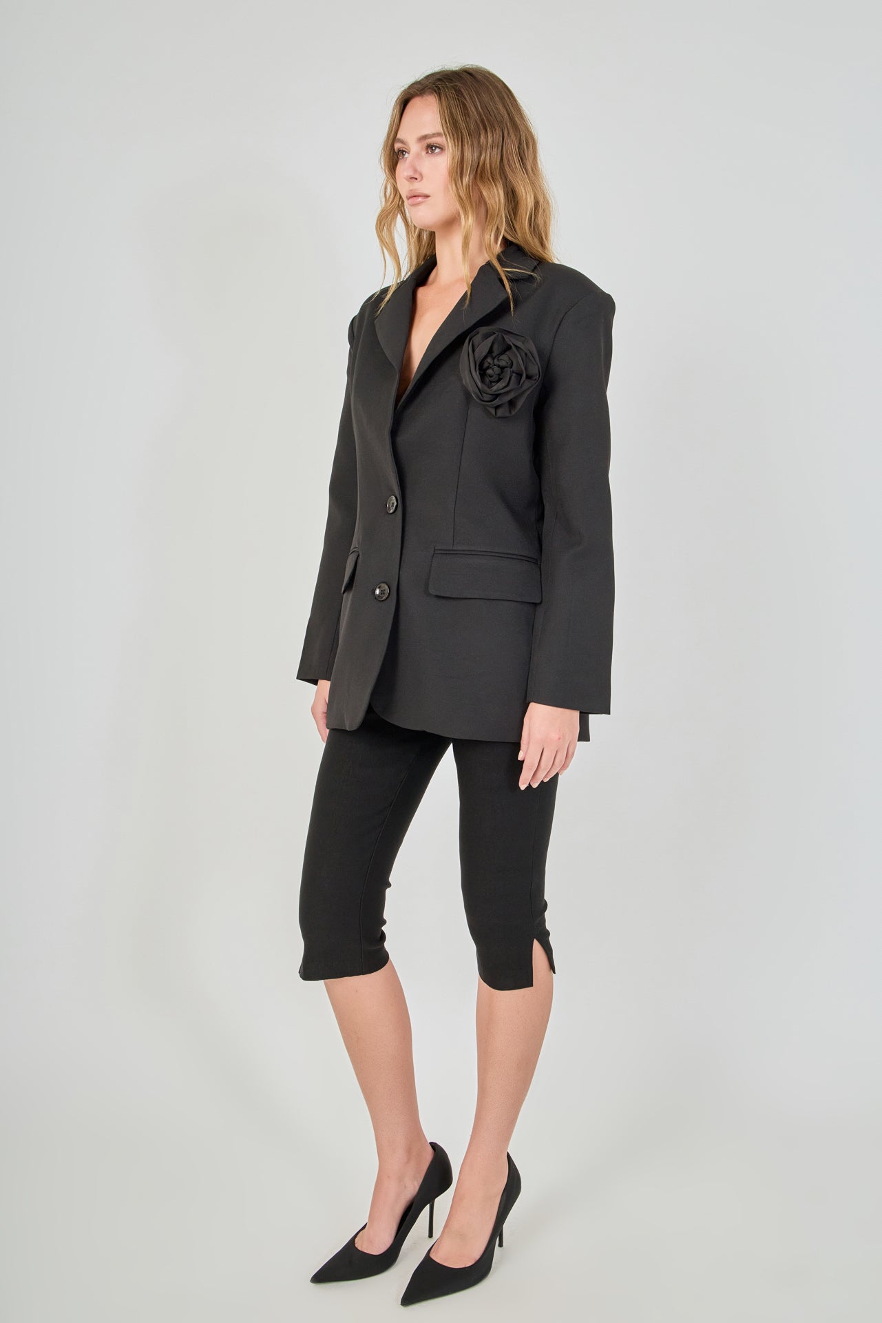 Side view of a black tailored blazer with structural shoulders and a straight hem, paired with fitted black capri pants showing the side-split detail.