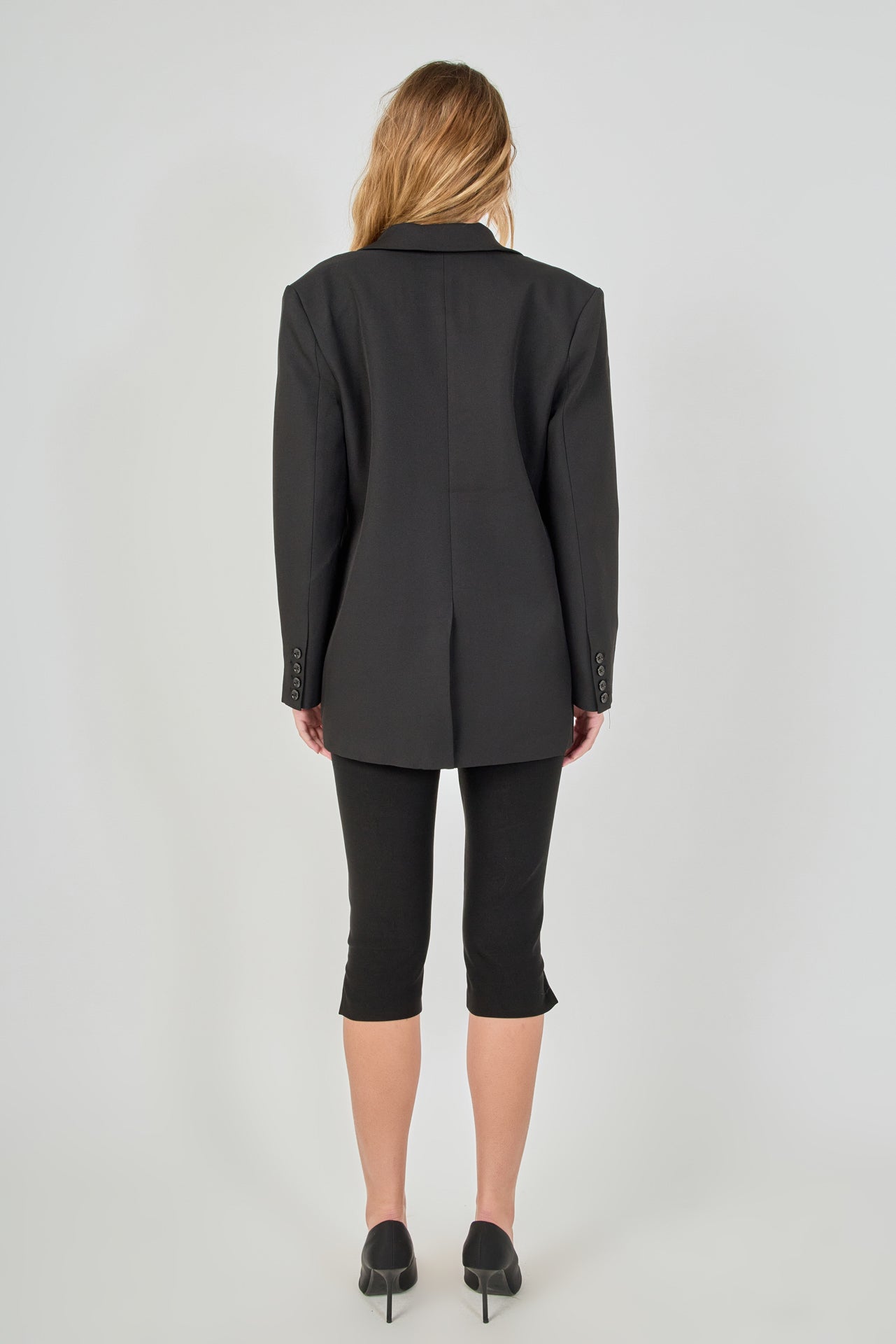 Back view of a black tailored blazer featuring a center back seam and a fitted silhouette, paired with fitted black capri pants.