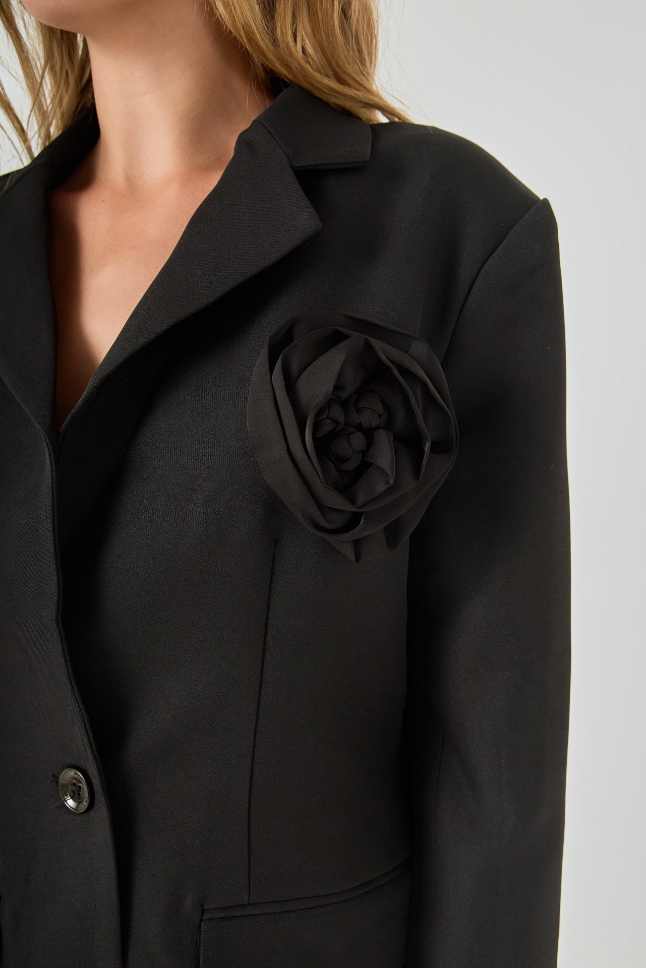 Close-up view of a black blazer highlighting the notch lapel, large intricate 3D rose appliqué, and textured fabric.