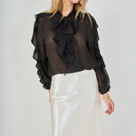 Mid-shot face-forward posed view of black sheer ruffled blouse featuring a dramatic tie-neck bow and cascading ruffles, paired with a high-waisted cream satin midi skirt.