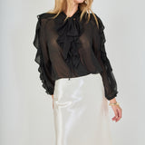 Mid-shot face-forward posed view of black sheer ruffled blouse featuring a dramatic tie-neck bow and cascading ruffles, paired with a high-waisted cream satin midi skirt.