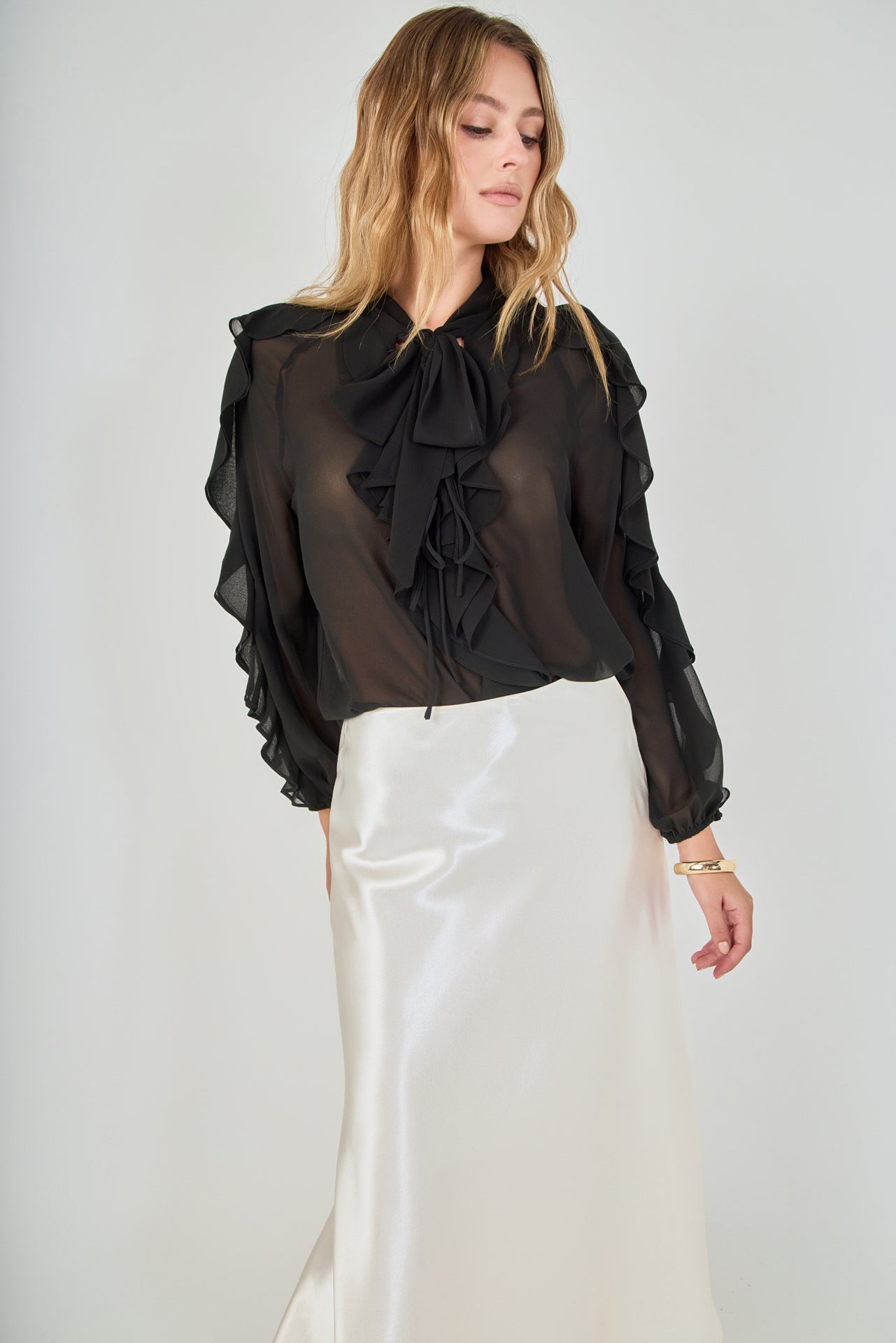 Mid-shot face-forward posed view of black sheer ruffled blouse featuring a dramatic tie-neck bow and cascading ruffles, paired with a high-waisted cream satin midi skirt.