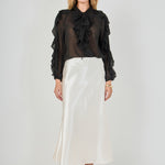 Full-length front view of a black sheer ruffled blouse featuring a tie-neck collar, cascading front ruffles, and long sheer sleeves with ruffle trim, styled with a cream satin midi skirt.