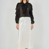 Full-length front view of a black sheer ruffled blouse featuring a tie-neck collar, cascading front ruffles, and long sheer sleeves with ruffle trim, styled with a cream satin midi skirt.