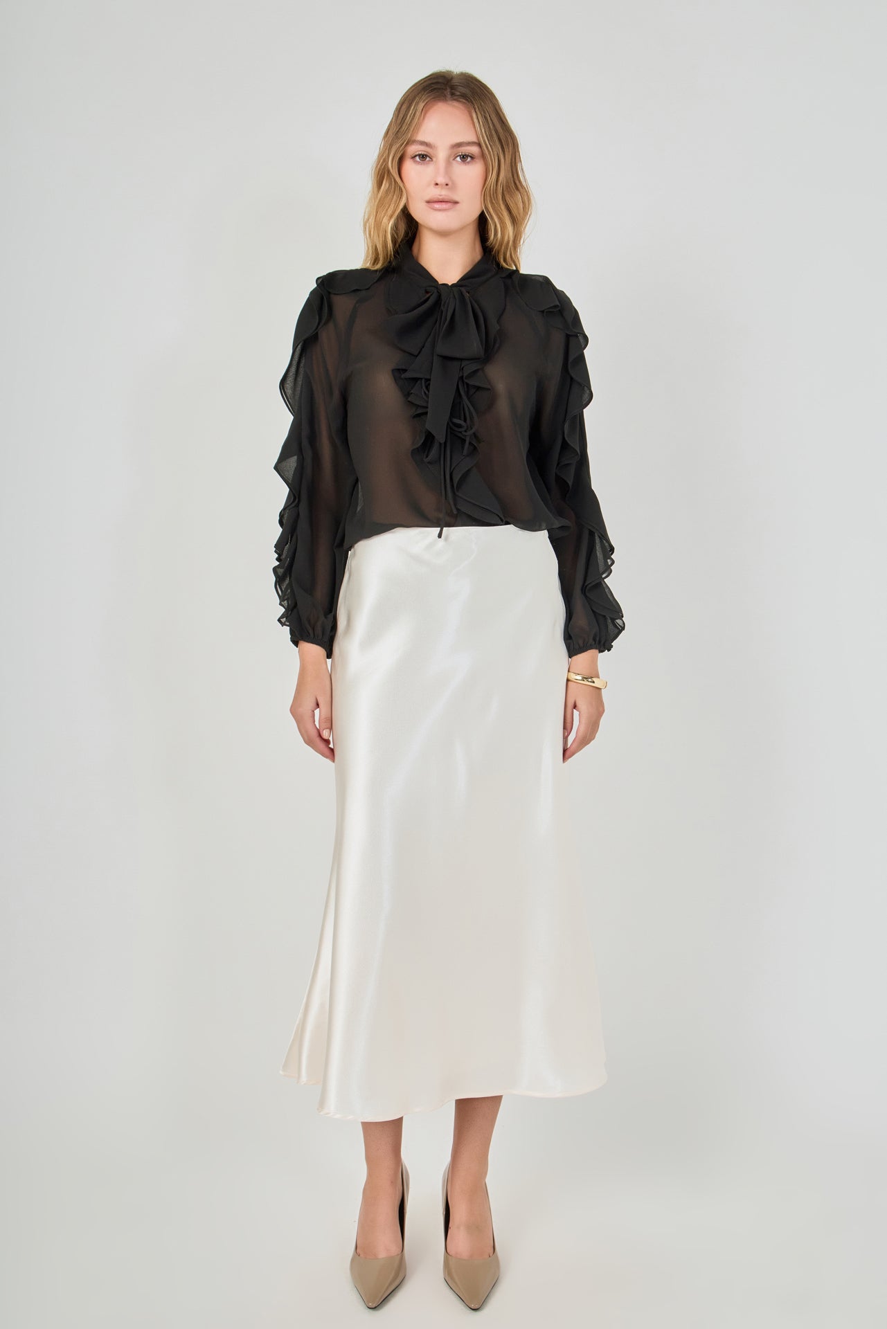 Full-length front view of a black sheer ruffled blouse featuring a tie-neck collar, cascading front ruffles, and long sheer sleeves with ruffle trim, styled with a cream satin midi skirt.