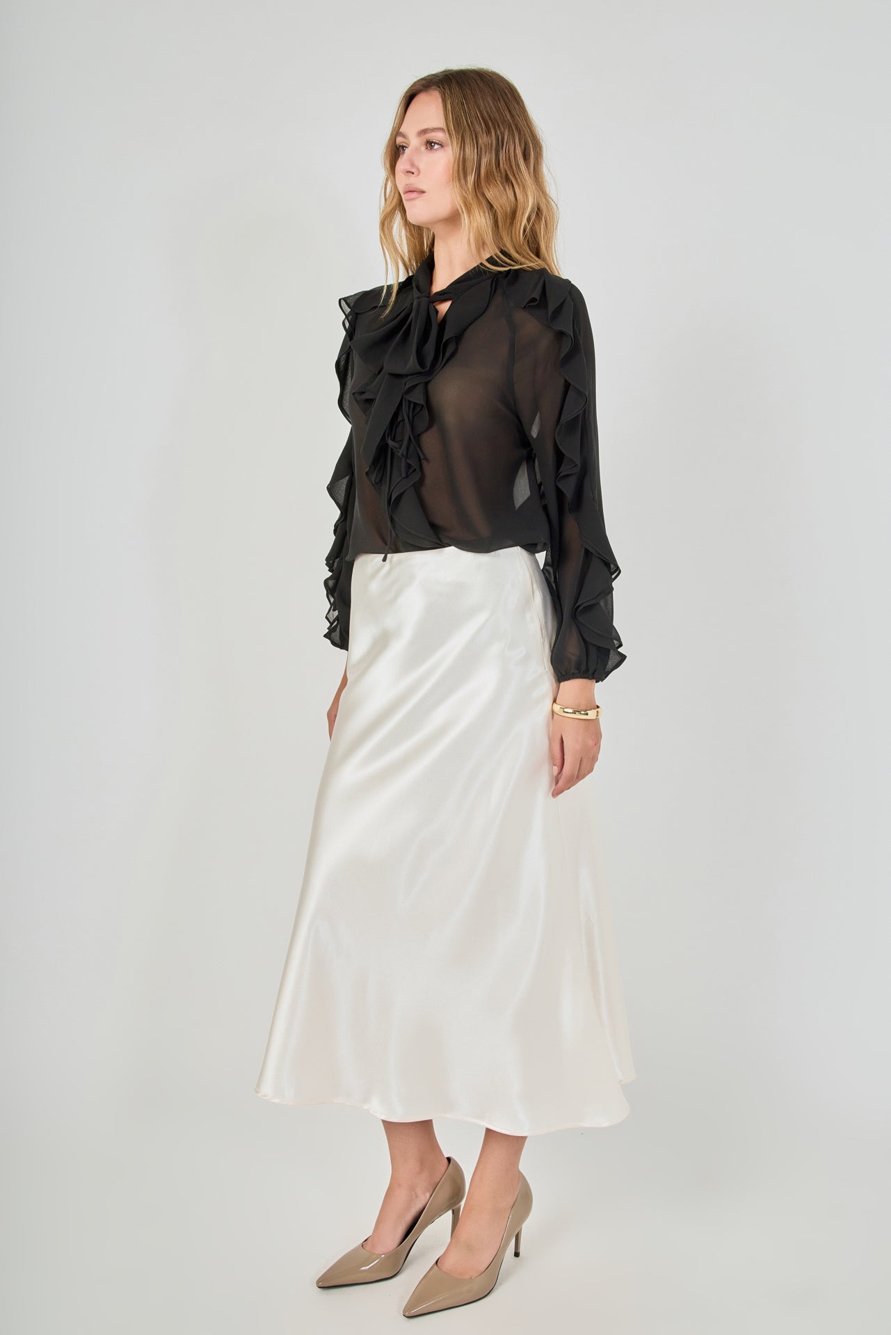 Side view of black sheer ruffled blouse featuring long sleeves with cascading outer ruffles, paired with a fluid cream satin midi skirt.