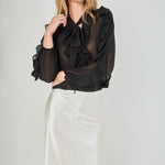 Mid-shot face-forward posed view of black sheer ruffled blouse featuring a dramatic tie-neck bow and cascading ruffles, paired with a high-waisted cream satin midi skirt.