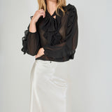 Mid-shot face-forward posed view of black sheer ruffled blouse featuring a dramatic tie-neck bow and cascading ruffles, paired with a high-waisted cream satin midi skirt.