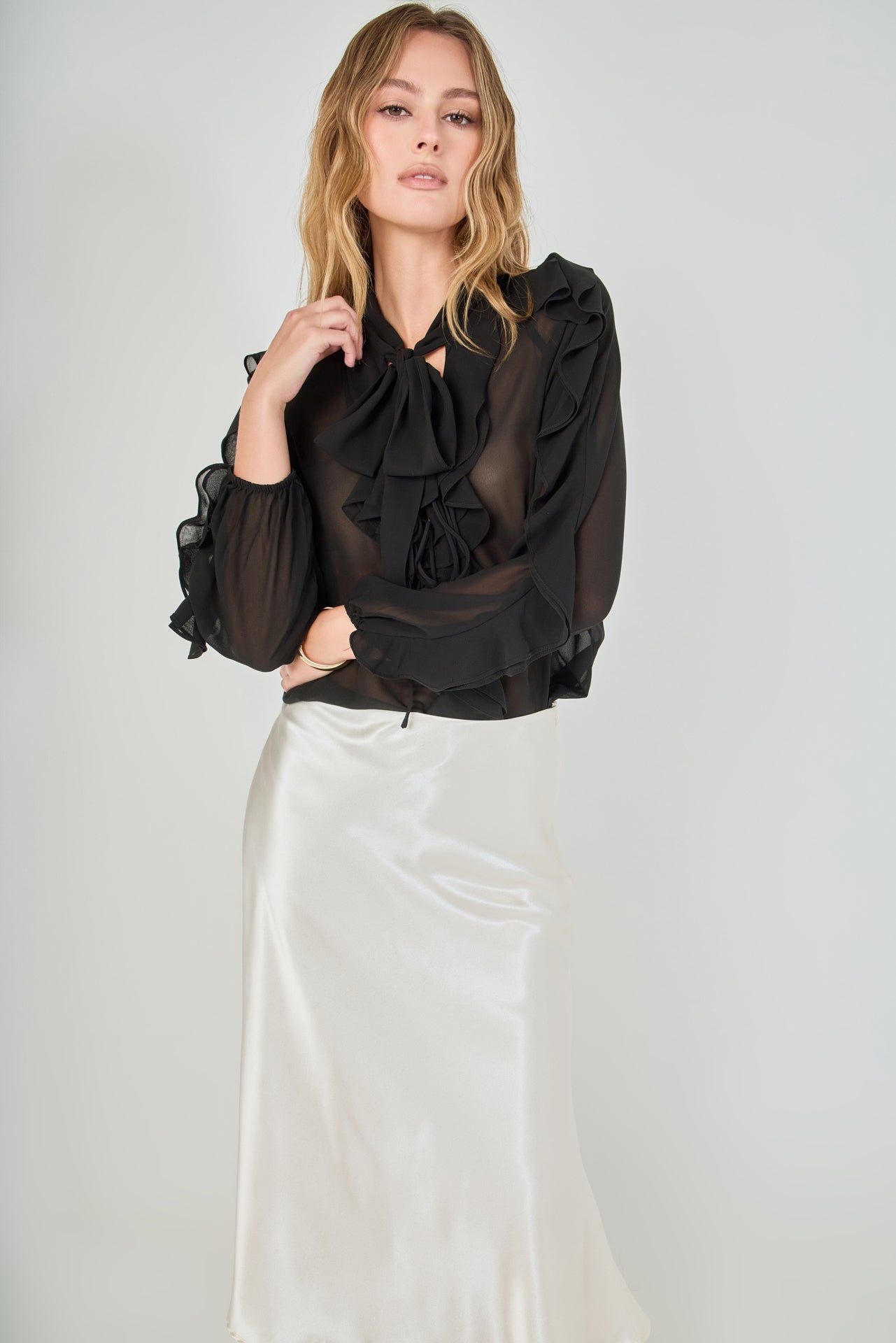 Mid-shot face-forward posed view of black sheer ruffled blouse featuring a dramatic tie-neck bow and cascading ruffles, paired with a high-waisted cream satin midi skirt.
