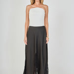 Full-length front view of a black maxi skirt featuring triangular sheer lace godet inserts and a scalloped eyelash lace hem, styled with a white ribbed strapless top.