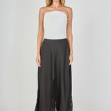 Full-length front view of a black maxi skirt featuring triangular sheer lace godet inserts and a scalloped eyelash lace hem, styled with a white ribbed strapless top.