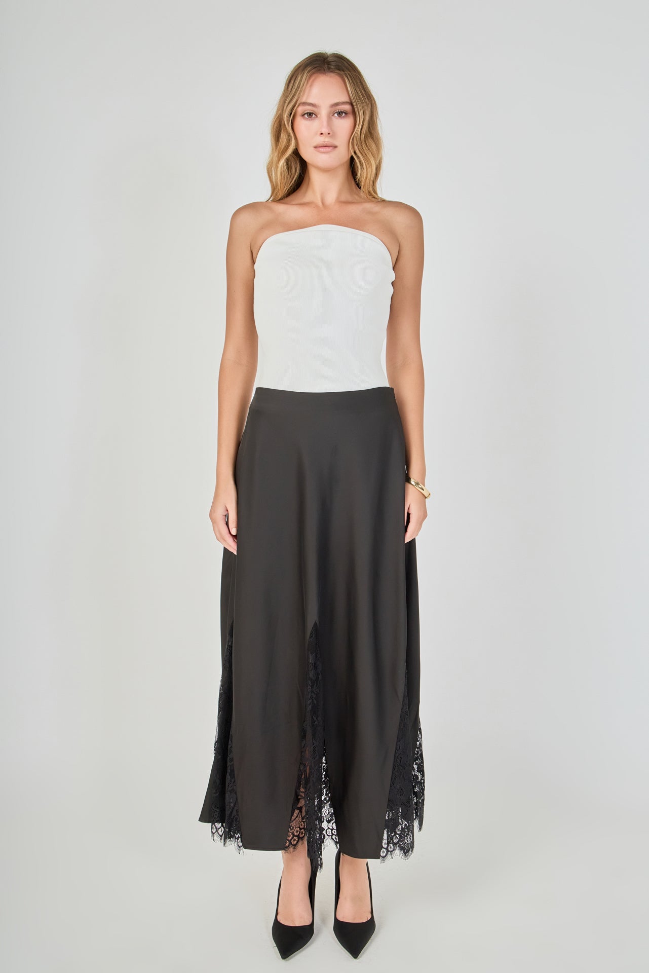 Full-length front view of a black maxi skirt featuring triangular sheer lace godet inserts and a scalloped eyelash lace hem, styled with a white ribbed strapless top.