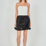 Full-length front view of a strapless mixed-media mini dress featuring a white ribbed knit tube bodice and a voluminous black 3D rosette sequined bubble mini skirt.