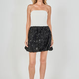 Full-length front view of a strapless mixed-media mini dress featuring a white ribbed knit tube bodice and a voluminous black 3D rosette sequined bubble mini skirt.