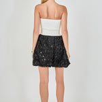 Back view of a strapless mixed-media mini dress featuring a smooth white ribbed back bodice and a black 3D rosette sequined mini hem.