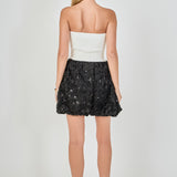 Back view of a strapless mixed-media mini dress featuring a smooth white ribbed back bodice and a black 3D rosette sequined mini hem.