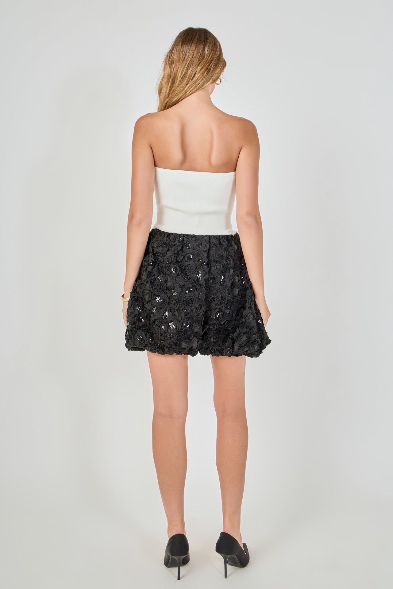 Back view of a strapless mixed-media mini dress featuring a smooth white ribbed back bodice and a black 3D rosette sequined mini hem.