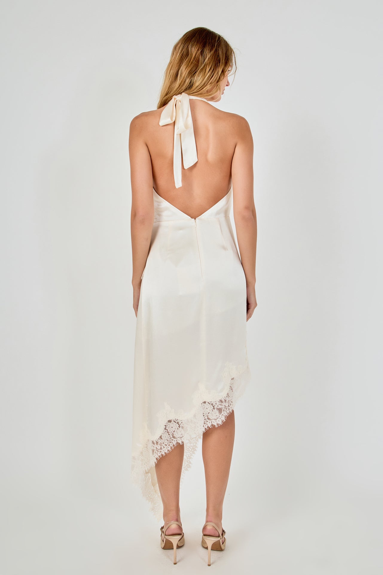 Straight-on back view of a champagne satin dress displaying a self-tie halter neck, open low back, and asymmetrical lace skirt hem.
