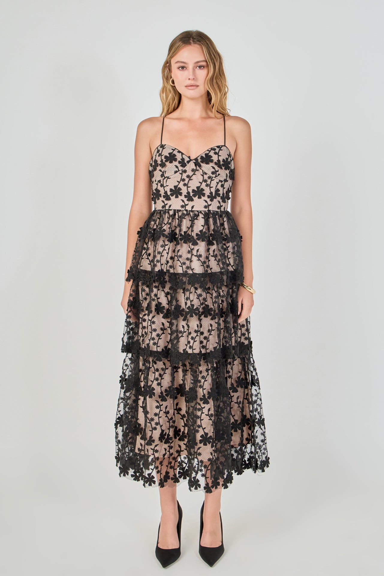 Full-length front view of a black floral embroidered tiered midi dress featuring a sweetheart neckline, thin spaghetti straps, sheer mesh overlay with black floral embroidery, contrast beige lining, and a tiered ruffled skirt.